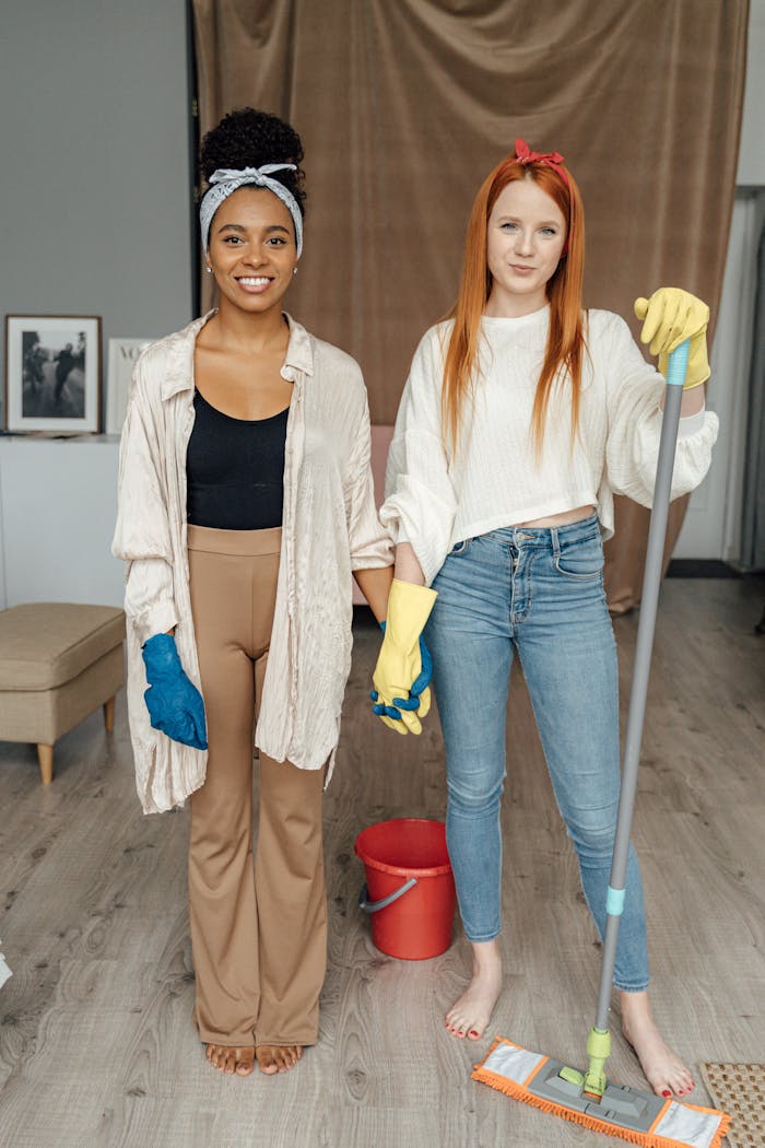 hero-img-02 Two women smiling and holding hands while cleaning indoors, symbolizing teamwork.